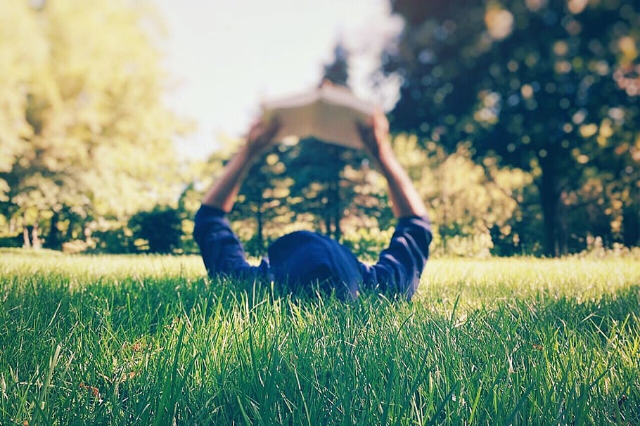 person in blue hoodie holding book while lying on green grass during daytime, HD wallpaper