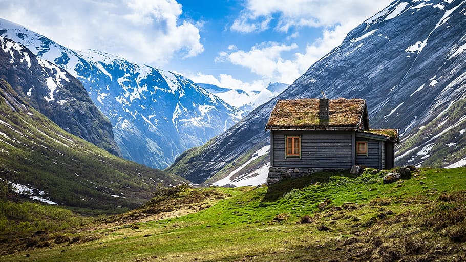 photo of brown wooden house on top of green hill near snow mountains during day time