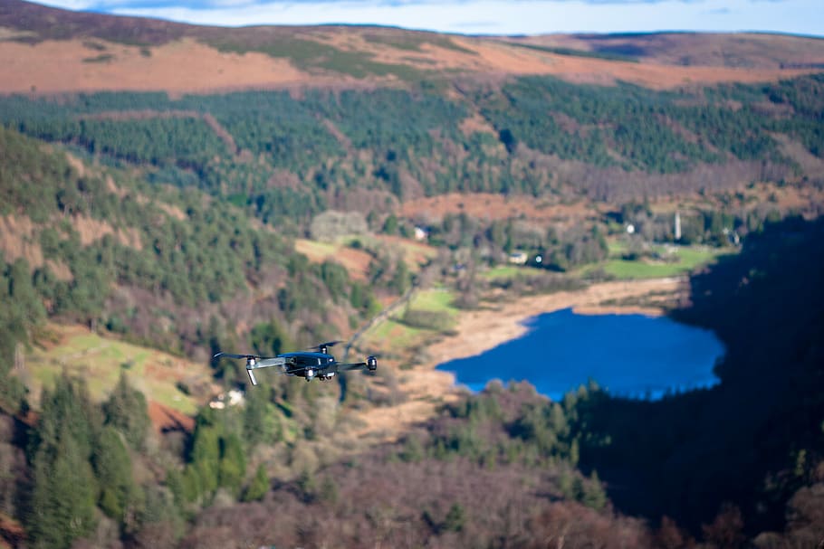 high-angle tilt-shift lens photography of black quadcopter drone hovering above forested valley during daytime, bird's-eye view photography of mountains with body of water HD wallpaper