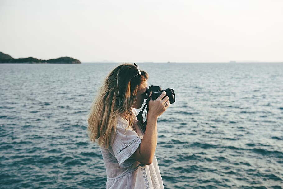 woman wearing white shirt holding camera taking photo of ocean during daytime HD wallpaper