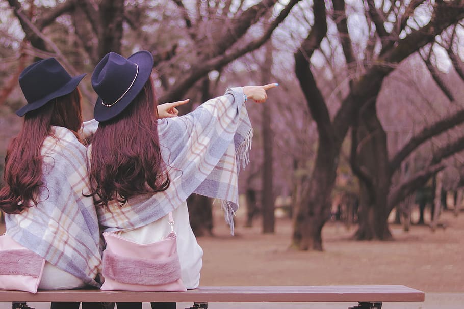 selective focus photography of two women wearing blue fedora hats pointing at the right side sitting in brown wooden bench chair during daytime, HD wallpaper