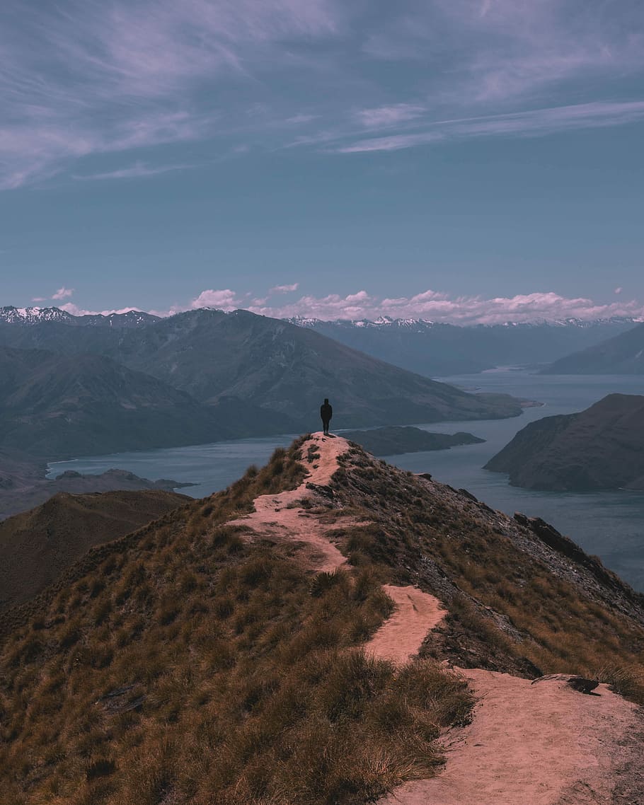 person standing on cliff near mountain under cloudy sky during daytime, man standing on green mountain HD wallpaper