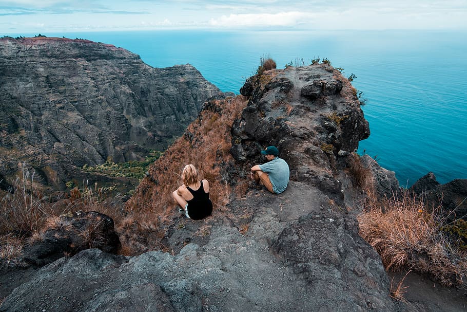 man and woman seating on mountain during day time, two person sitting on hill over body of water