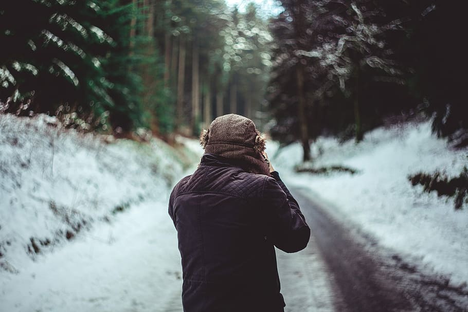 man in brown jacket standing between trees, man walking on snowy path HD wallpaper