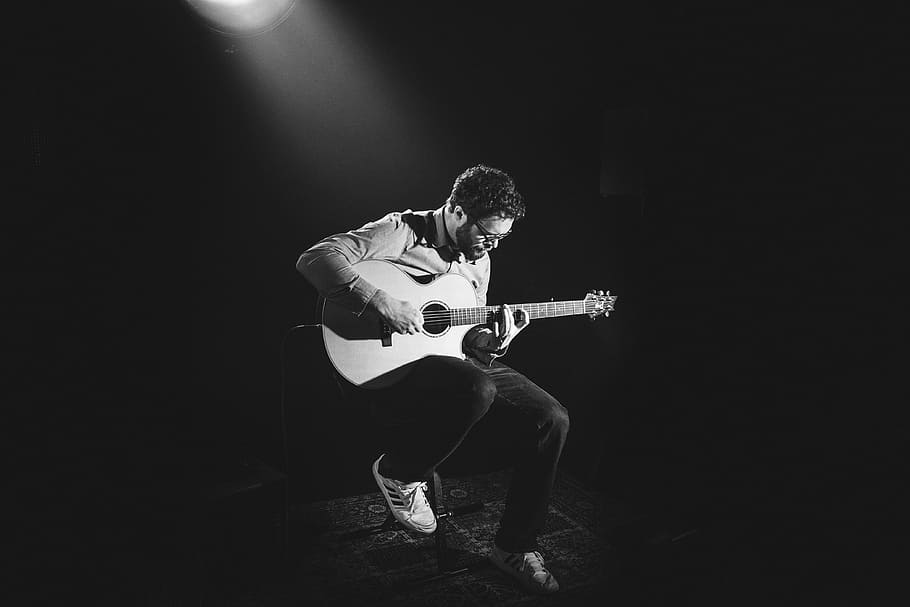 grayscale photo of man playing guitar on stage, grayscale photography og man sitting on chair while playing acoustic guitar