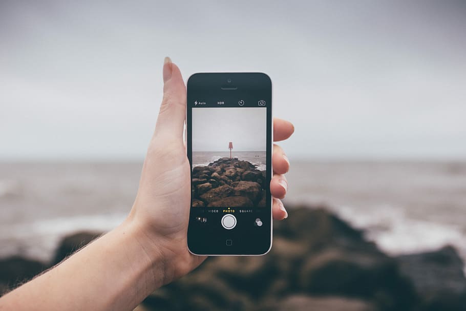 person holding black iPhone 4 while taking photo of sea shore during daytime HD wallpaper
