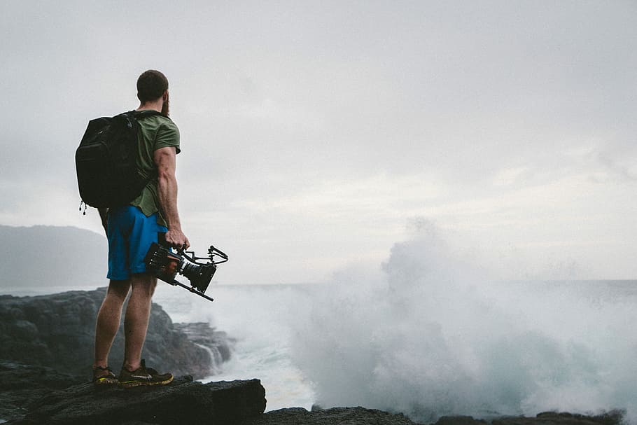man in green shirt standing near clouds, man carrying a backpack and video recorder on rock formation near body of water HD wallpaper