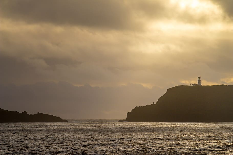 silhouette photo of lighthouse on mountain across body of water during golden hour HD wallpaper