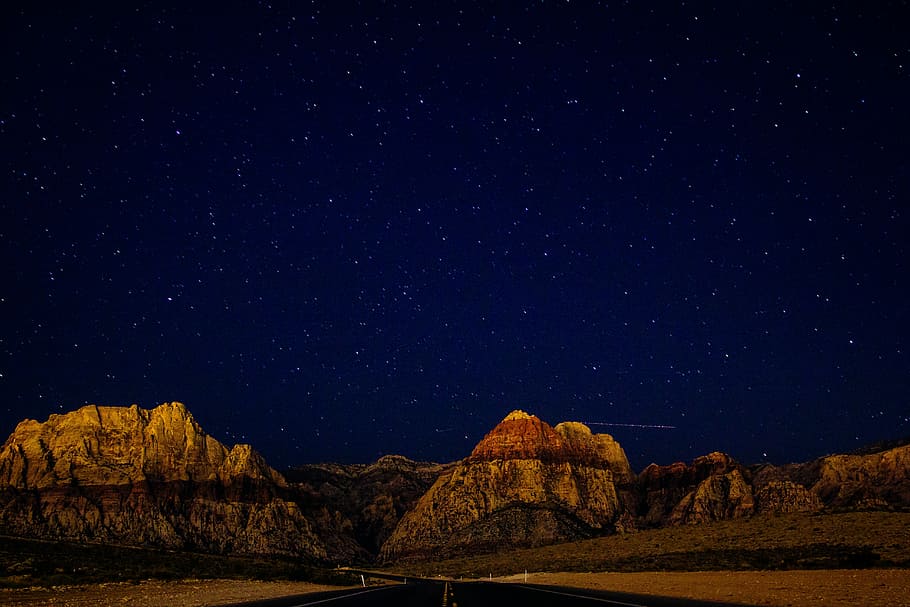 rocky mountain under star at night, empty highway overlooking brown rock mountain under stars at nighttime
