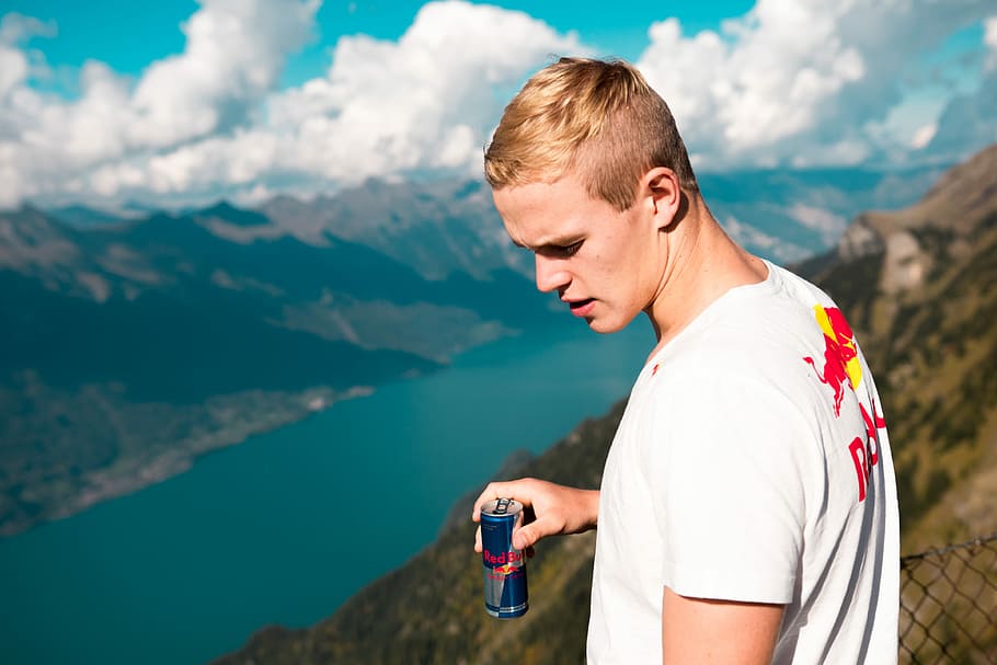 man wearing white and red crew-neck t-shirt holding Red Bull energy drink can standing on summit with chain link fence looking at body of water during daytime, selective focus photography of man holding Red Bull can