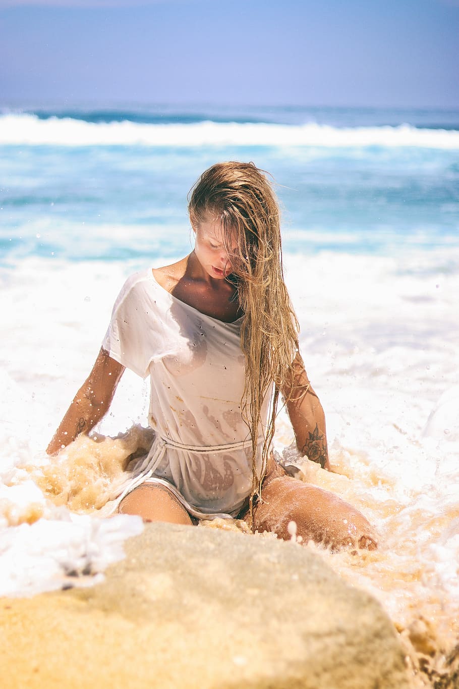 woman in white top sitting on sand near body of water, selective focus woman sitting on seashore during daytime