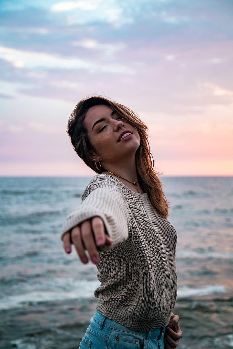 woman in grey sweater and blue denim bottoms reaching out arms and slightly looking up at beach under pink and blue sky, selective focus photography of woman standing near sea HD wallpaper
