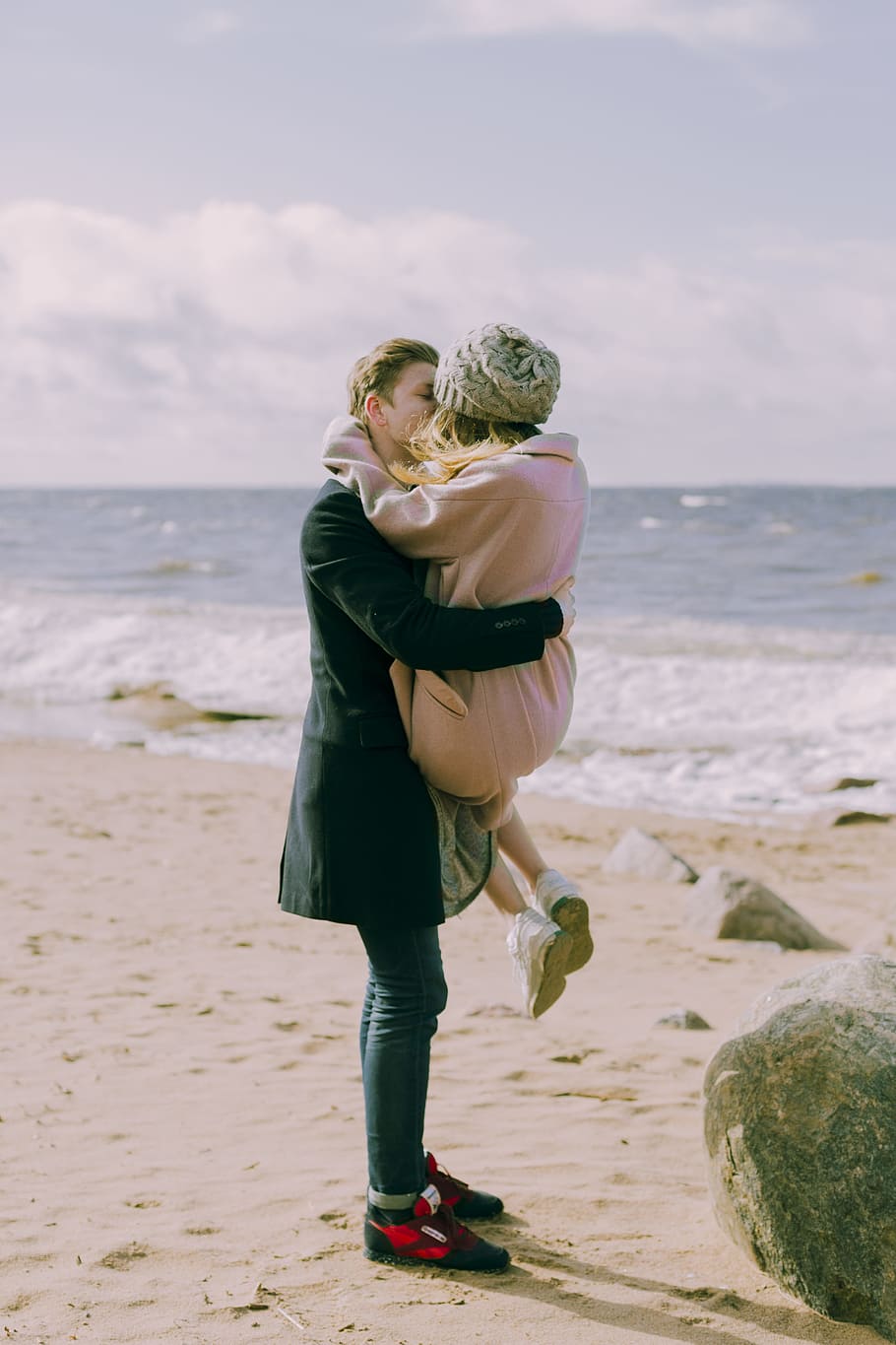Man in Black Overcoat and Blue Denim Jeans Kissing While Carrying a Woman in Pink Overcoat and Knit Cap on Shore at Daytime HD wallpaper