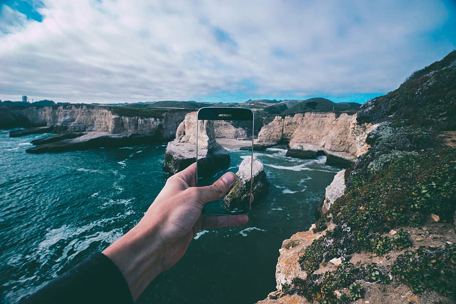 person holding black smartphone capturing the rock formation beyond body of water during daytime HD wallpaper