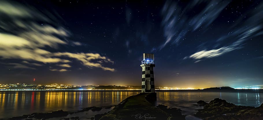 gray concrete under white cloudy sky during night time photo, white and black lighthouse near city buildings with light on selective focus photo HD wallpaper
