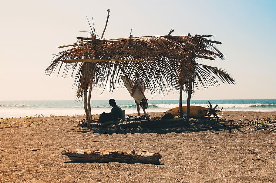 man reclining under hut on seashore, person sitting on brown canopy on seashore near man holding surfboard at daytime HD wallpaper