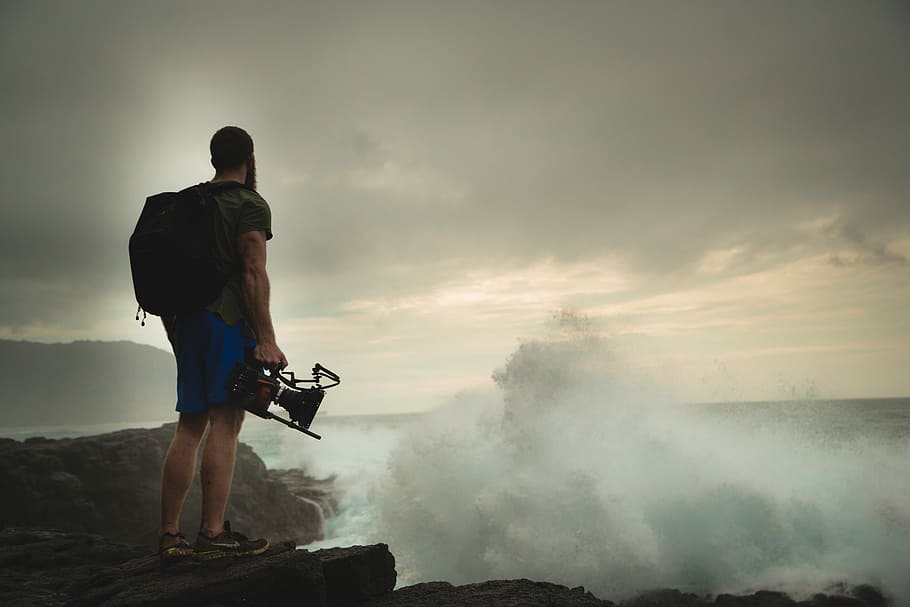 man standing on rock cliff near body of water, man standing on cliff while holding camera HD wallpaper