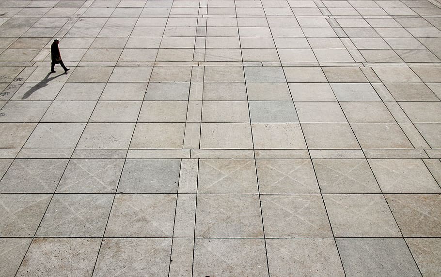 man walking on tiled ground at daytime, silhouette of person walking on gray concrete pavement high-angle photo