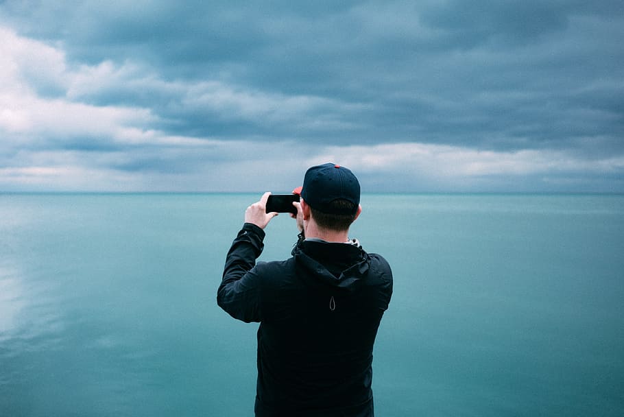 men's black hoodie in front of teal sea, person taking photo of blue sea and white clouds during daytime