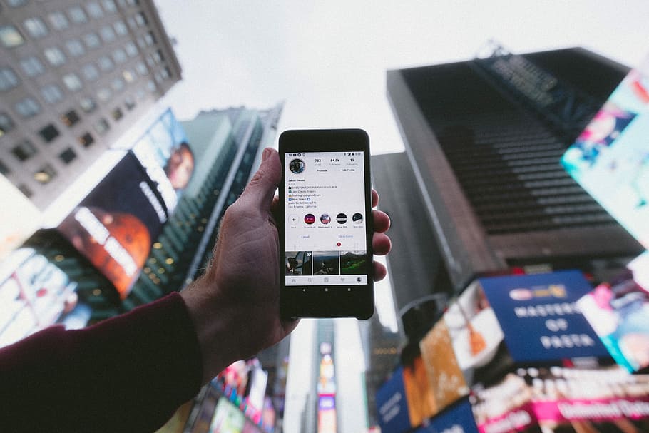 high angle photo of person holding turned on smartphone with tall buildings background, person holding smartphone showing instagram post HD wallpaper