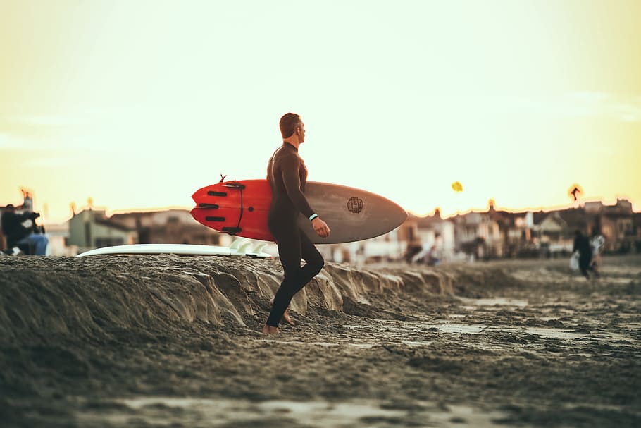person carrying surfboard on mud during daytime, man carrying red and gray surfboard while walking on the beach HD wallpaper