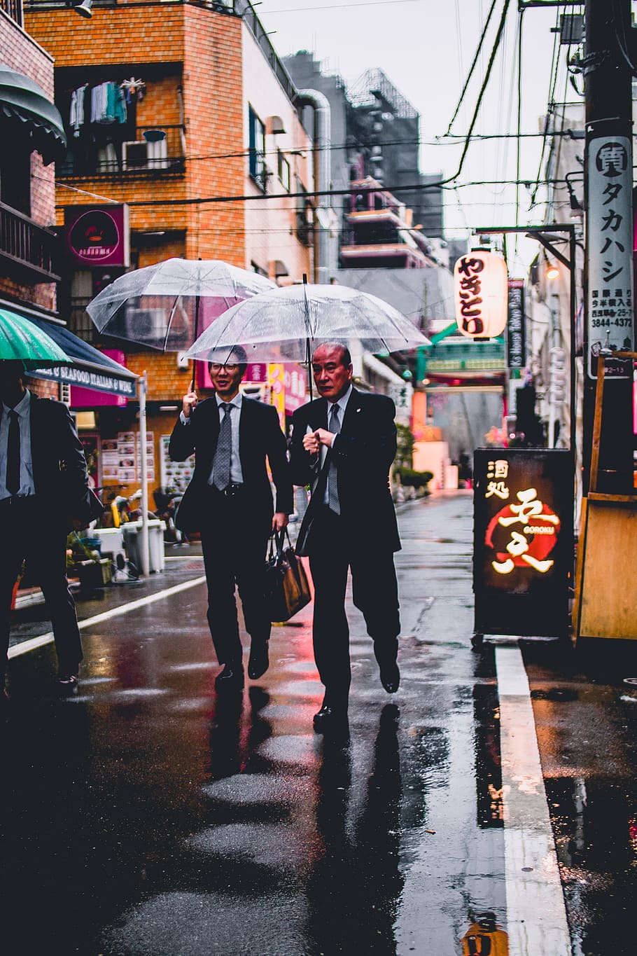 two men in black suit holding transparent umbrellas walking in the street, three man wearing black suits holding umbrella walking on street