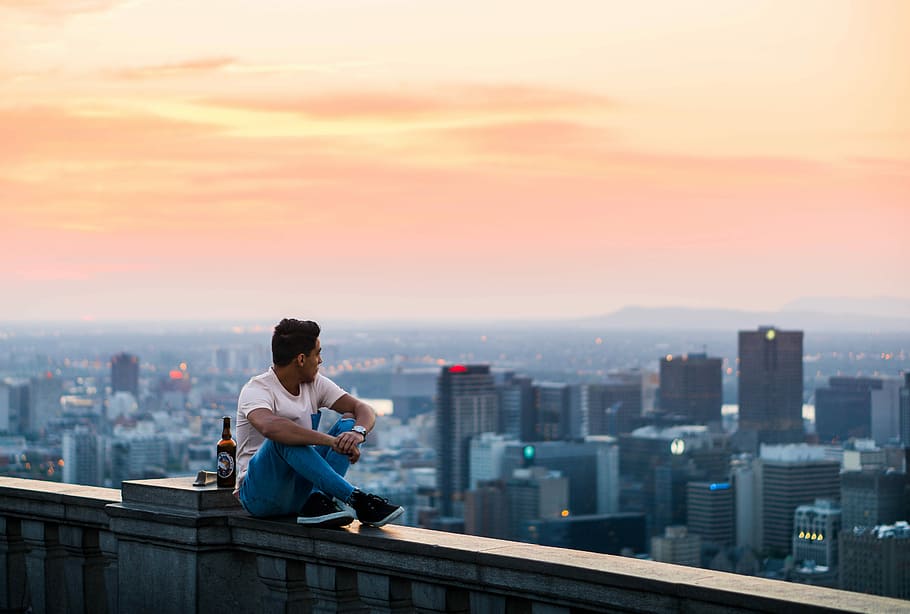 man seating beside buildings, man sitting on edge of building beside bottle watching high rise buildings during golden hour