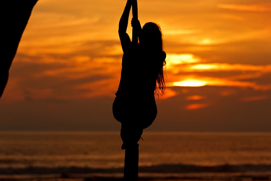 silhouette photo of a man holding rope on seashore during sunset, silhouette of woman on pole bar near body of water