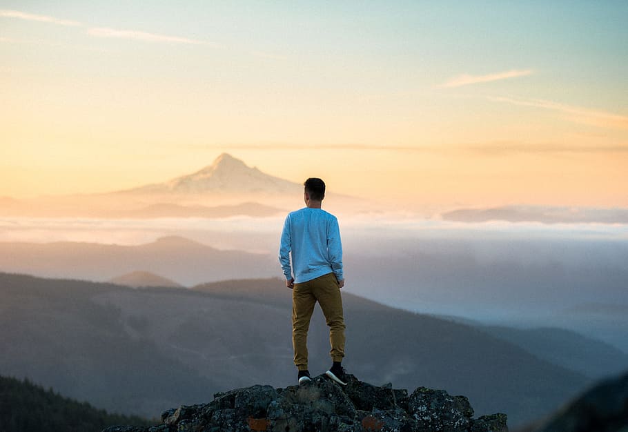 man standing on top of mountain, man in blue long-sleeved shirt standing on mountain during daytime