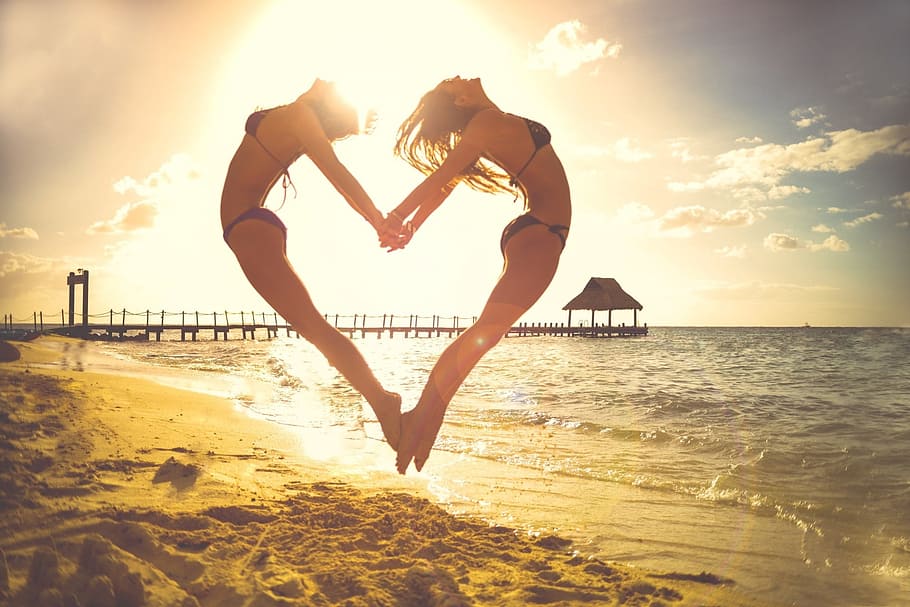 two women jumping while forming heart shape beside seashore during daytime, HD wallpaper
