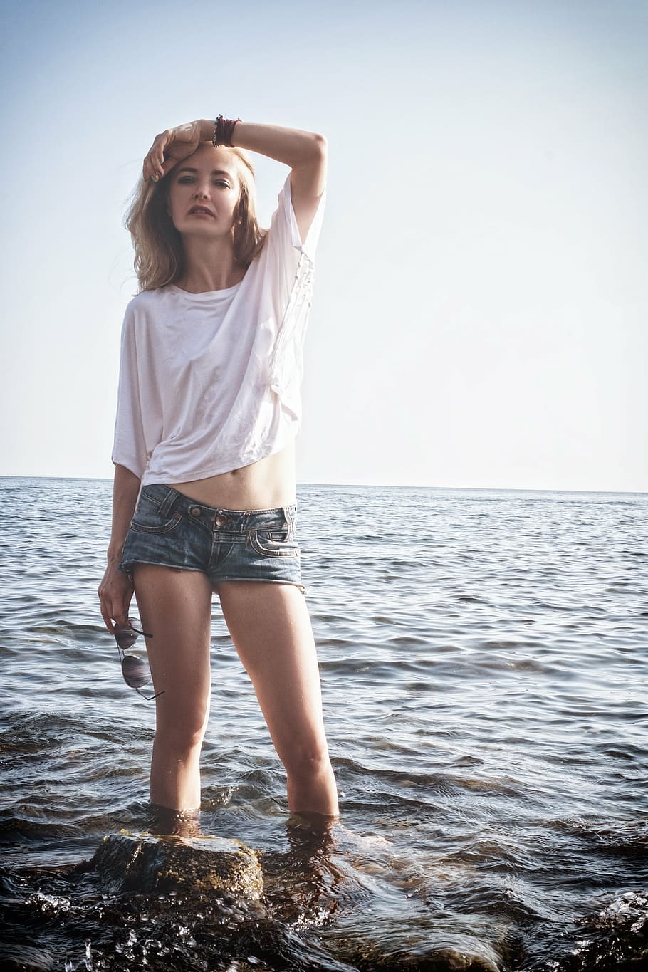 woman wearing white shirt and blue denim short shorts standing near rock on seashore at daytime, HD wallpaper