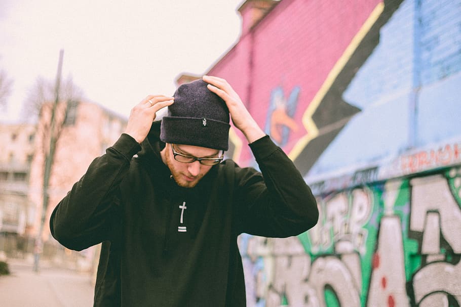 man touching his cap while walking, man in black pullover hooded jacket wearing knit cap standing beside wall during daytime HD wallpaper