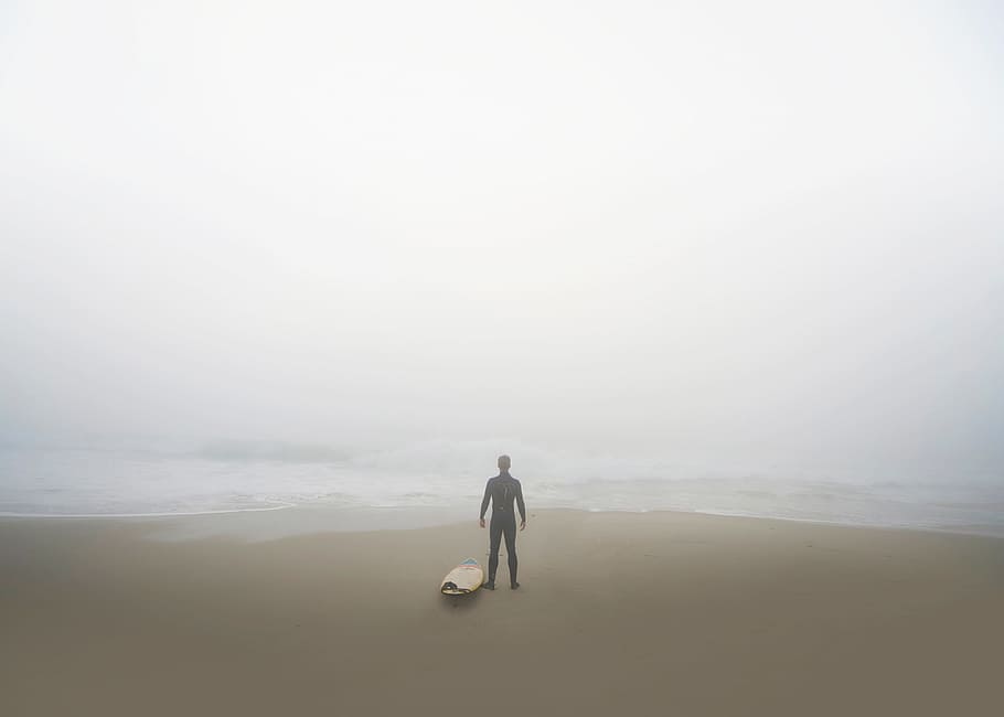 man standing beside surfboard on seashore, silhouette of person standing facing body of water near surfboard, HD wallpaper