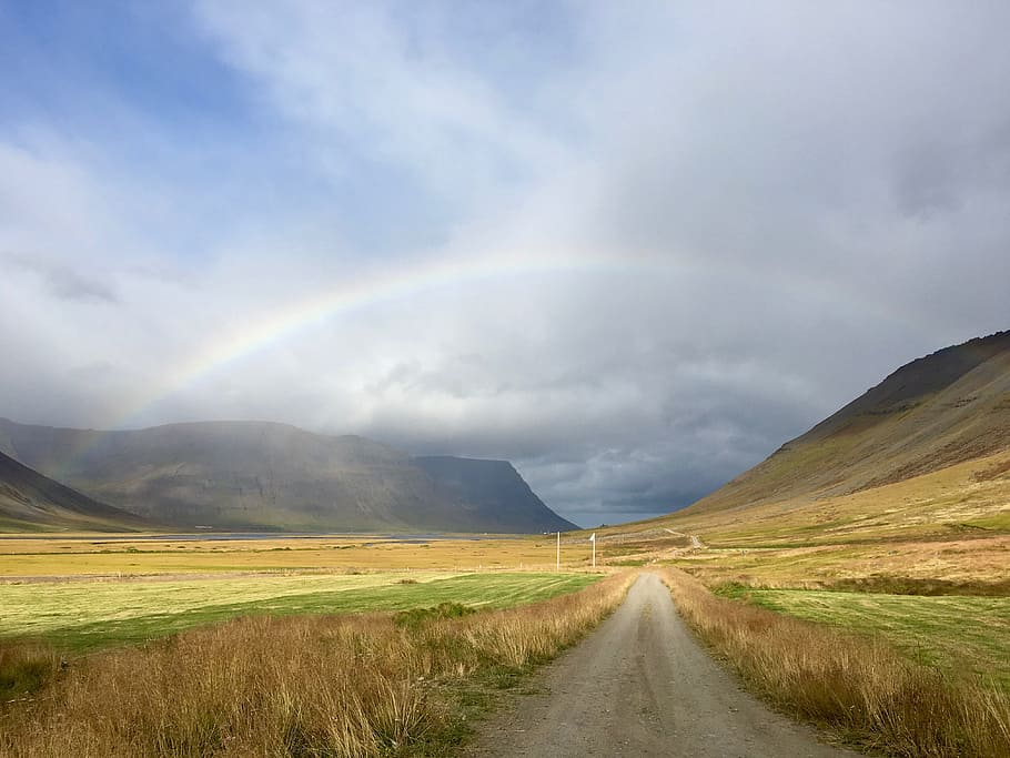 dirt path between brown and green field leading to a rainbow under blue and white cloudy sky, gray road between green grass near mountain under gray clouds with rainbow at daytime