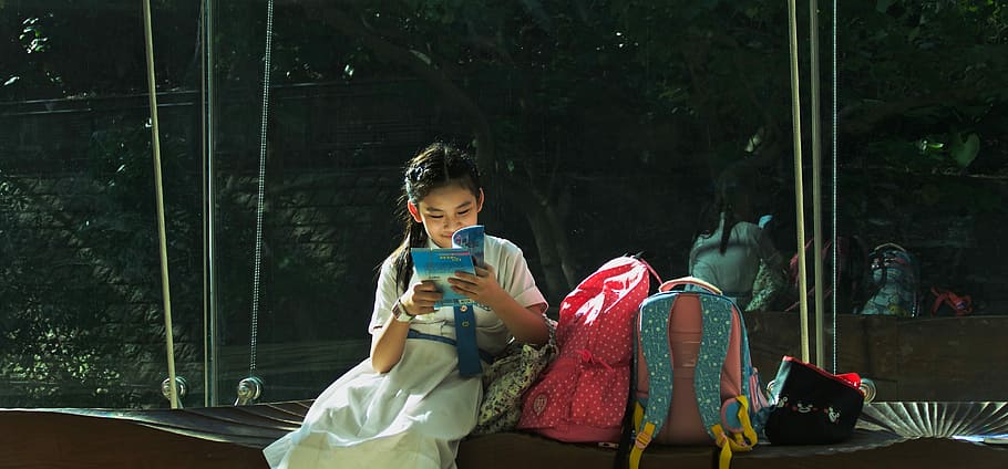 girl reading book, girl wearing white dress reading book beside two backpacks during daytime HD wallpaper