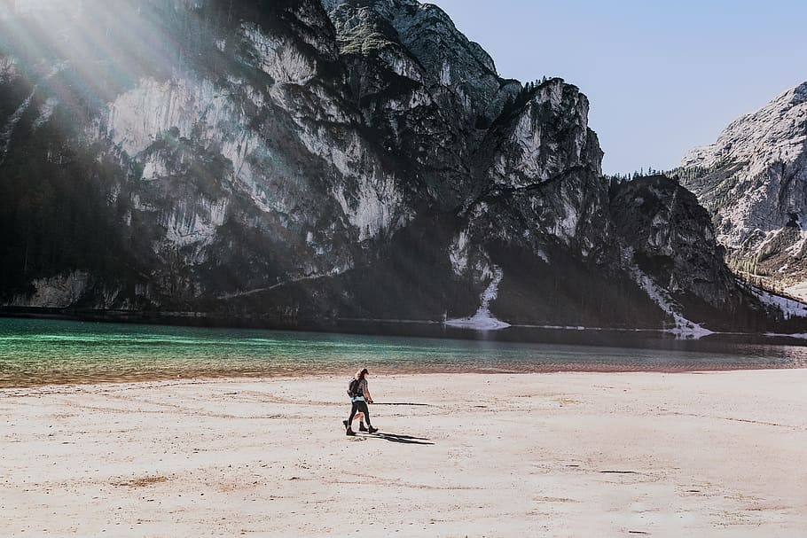 two people walks on beach, two person walking beside river and mountain at daytime HD wallpaper