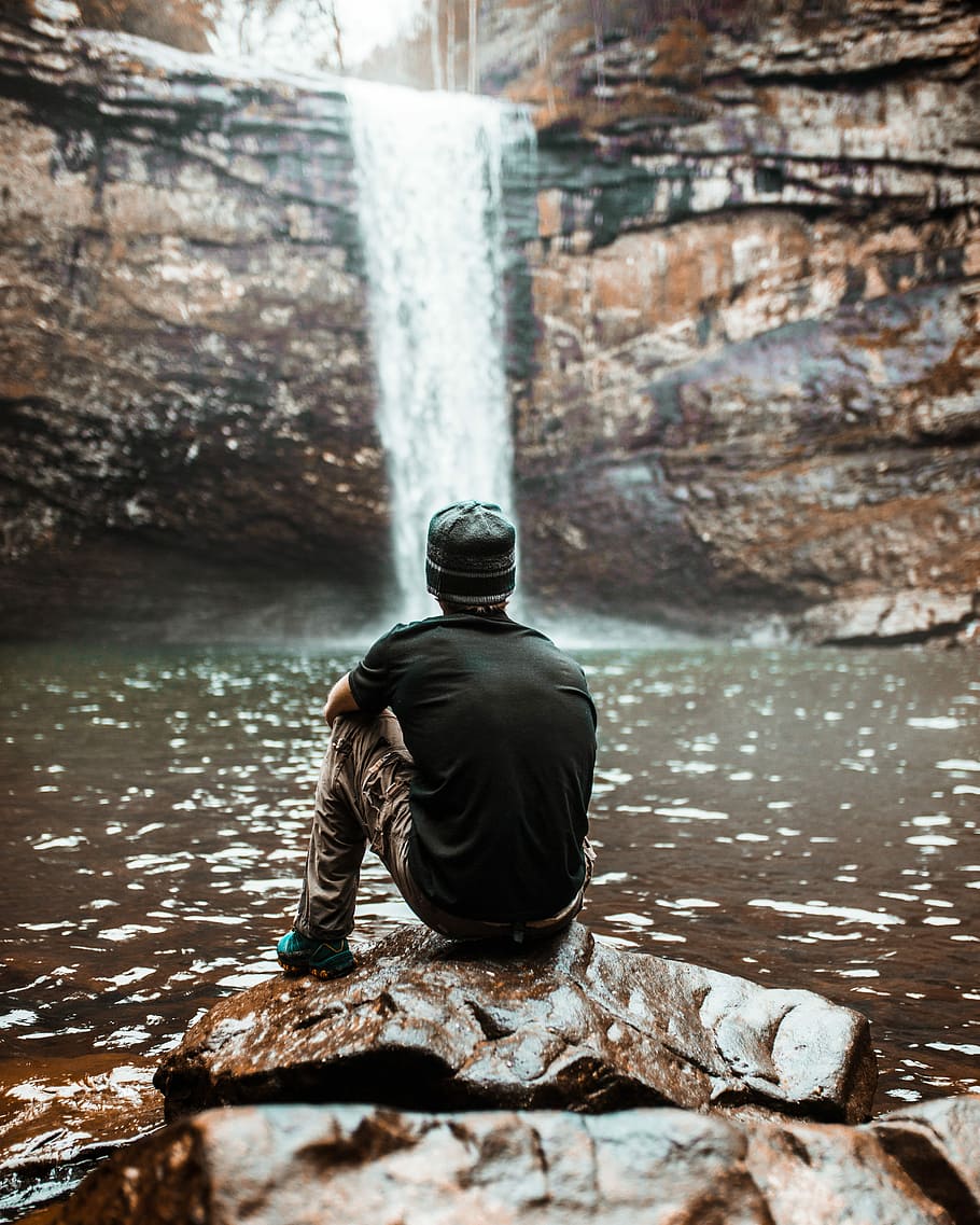 man sitting on brown rock near waterfalls at day time, man sitting on rock looking at waterfalls HD wallpaper
