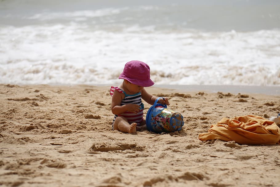 baby wearing purple hat playing sand near ocean at daytime, child playing