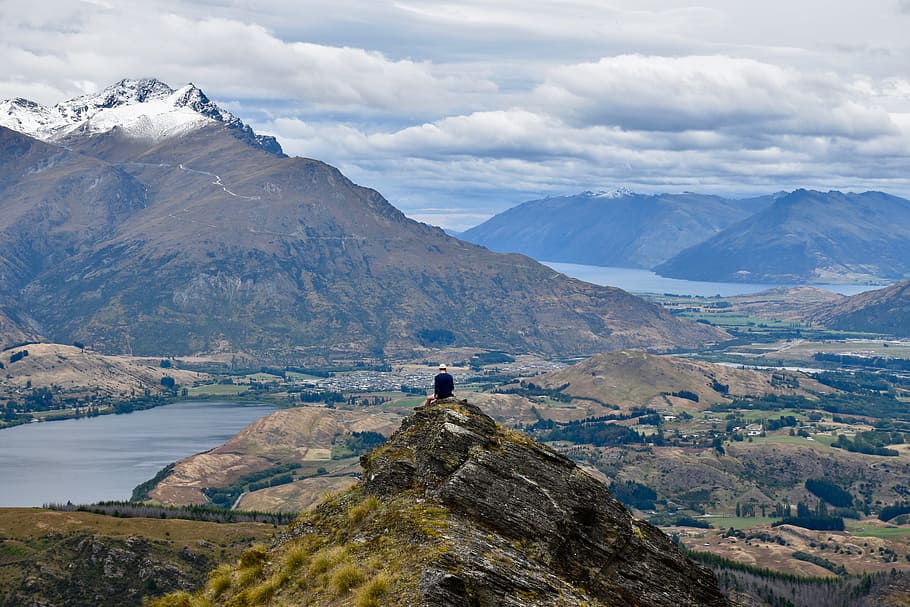 person standing on hill facing mountain and body of water under white and blue sky, person sitting on top of mountain's peak HD wallpaper