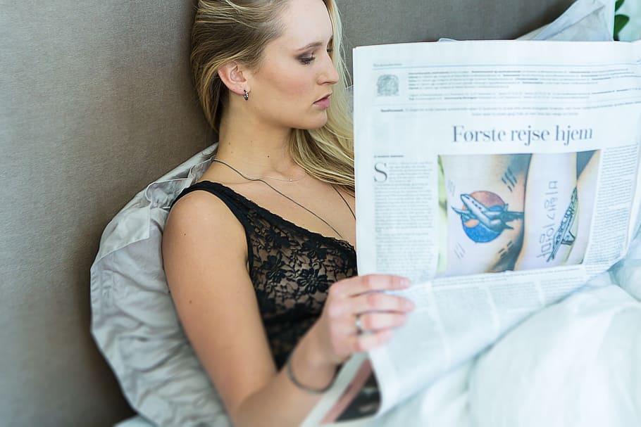 woman wearing black chemise reading newspaper, woman reading newspaper while leaning on gray wall