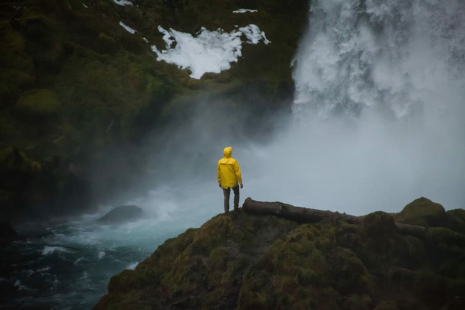 person in yellow coat standing near waterfalls during daytime, person standing on brown rock formation in front of waterfalls HD wallpaper
