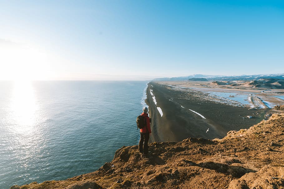man standing on cliff facing sea, person standing on cliff viewing sea under white and blue sky during daytime HD wallpaper
