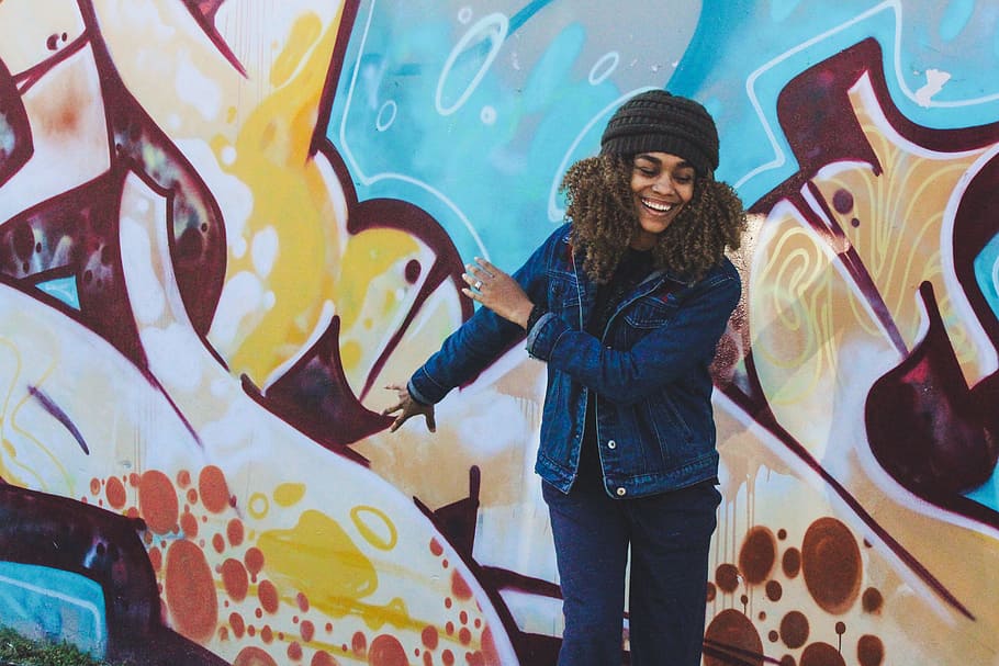 woman wearing knit hat and swing her arms, woman wearing blue denim jacket and blue jeans near graffiti wall