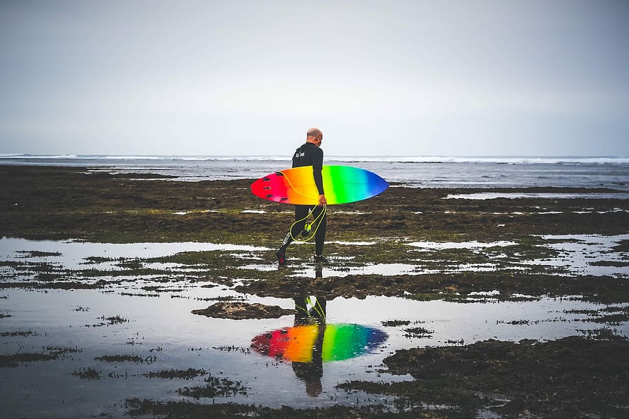 man in black wetsuit carrying red, yellow, blue, and green surfboard, man carrying multicolored surfboard walking on beach HD wallpaper