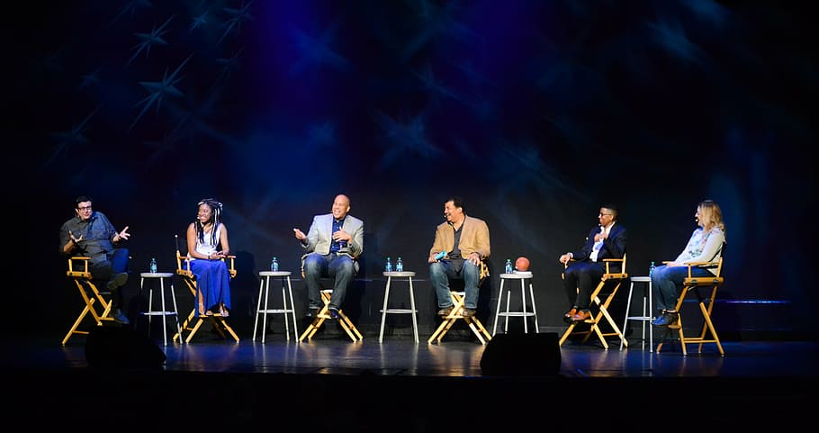 six people sitting on chairs on stage, talking, and share the