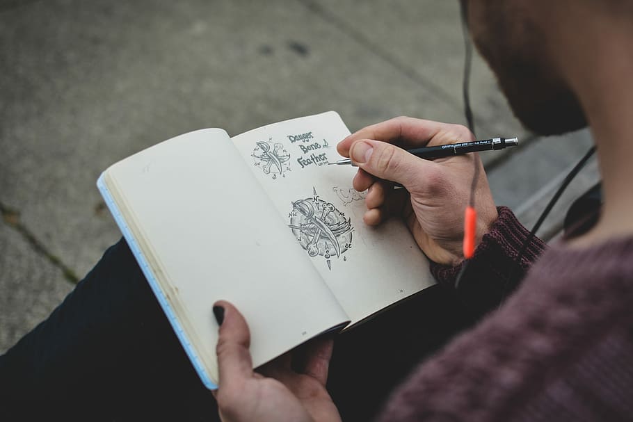 shallow focus photography of person drawing, person sits holding book and pen, HD wallpaper