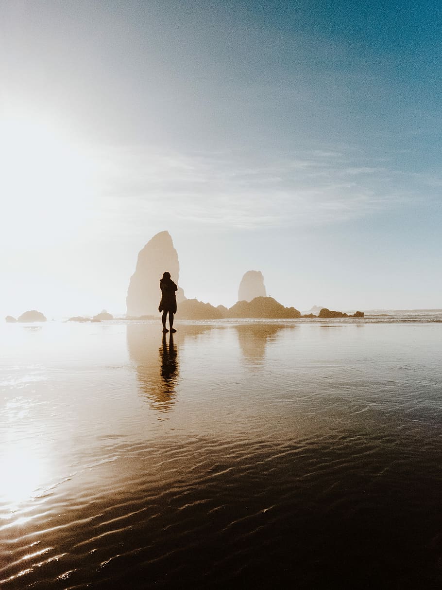 person standing in shallow body of water near boulders, silhouette of person standing on sea at daytime HD wallpaper