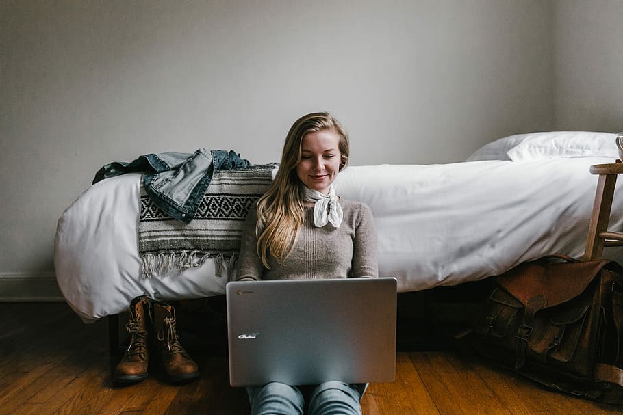 woman sitting beside a bed while using a laptop, photo of woman using silver laptop computer