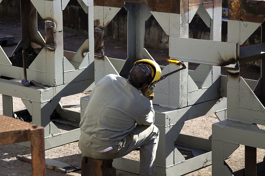 man sitting on brick holding acetylene and boding metal sheet on white frame at daytime HD wallpaper