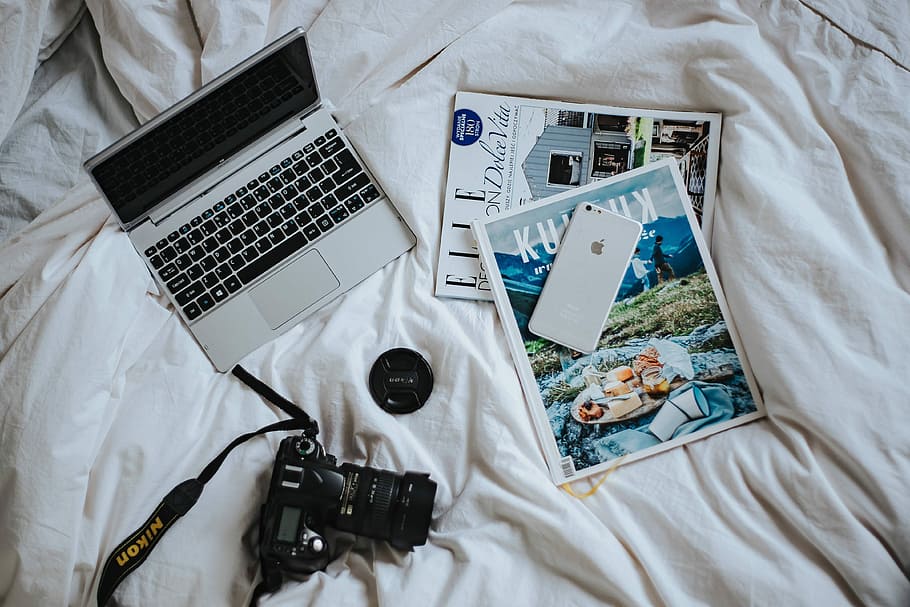 Silver laptop, a camera, magazines and other items on white bed sheets HD wallpaper